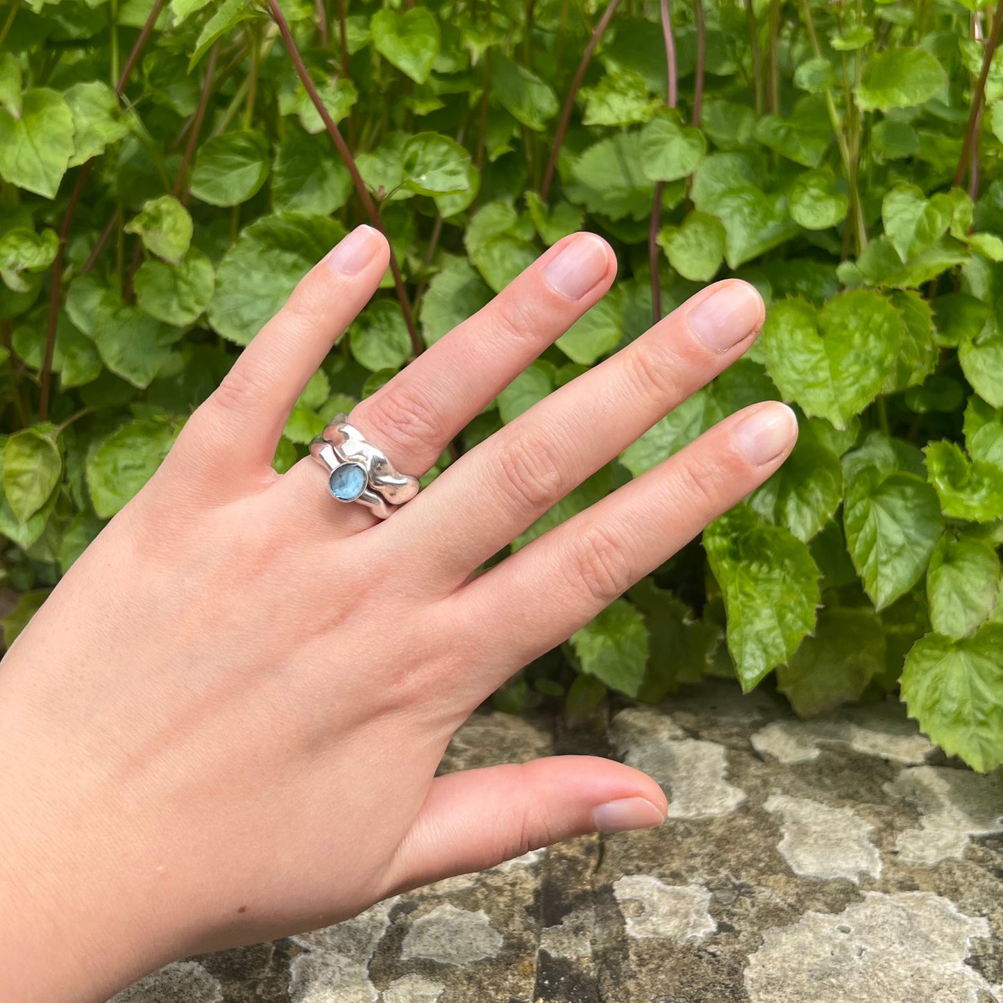 The silver wave ring with a topaz set bubble ring on a hand with a rock and leaf background.