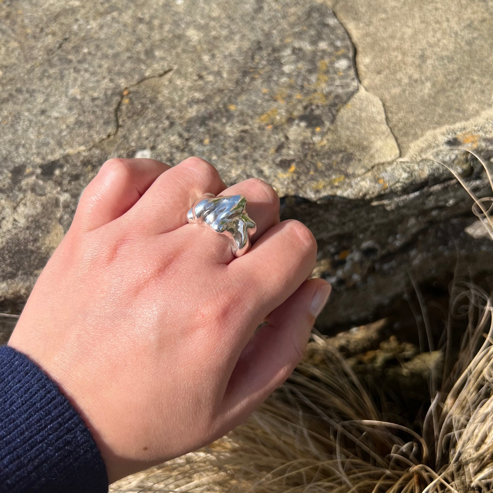 The Craggy ring from the side. A chunky statement ring handmade in sterling silver which is photographed on a hand. The background is of a rock.