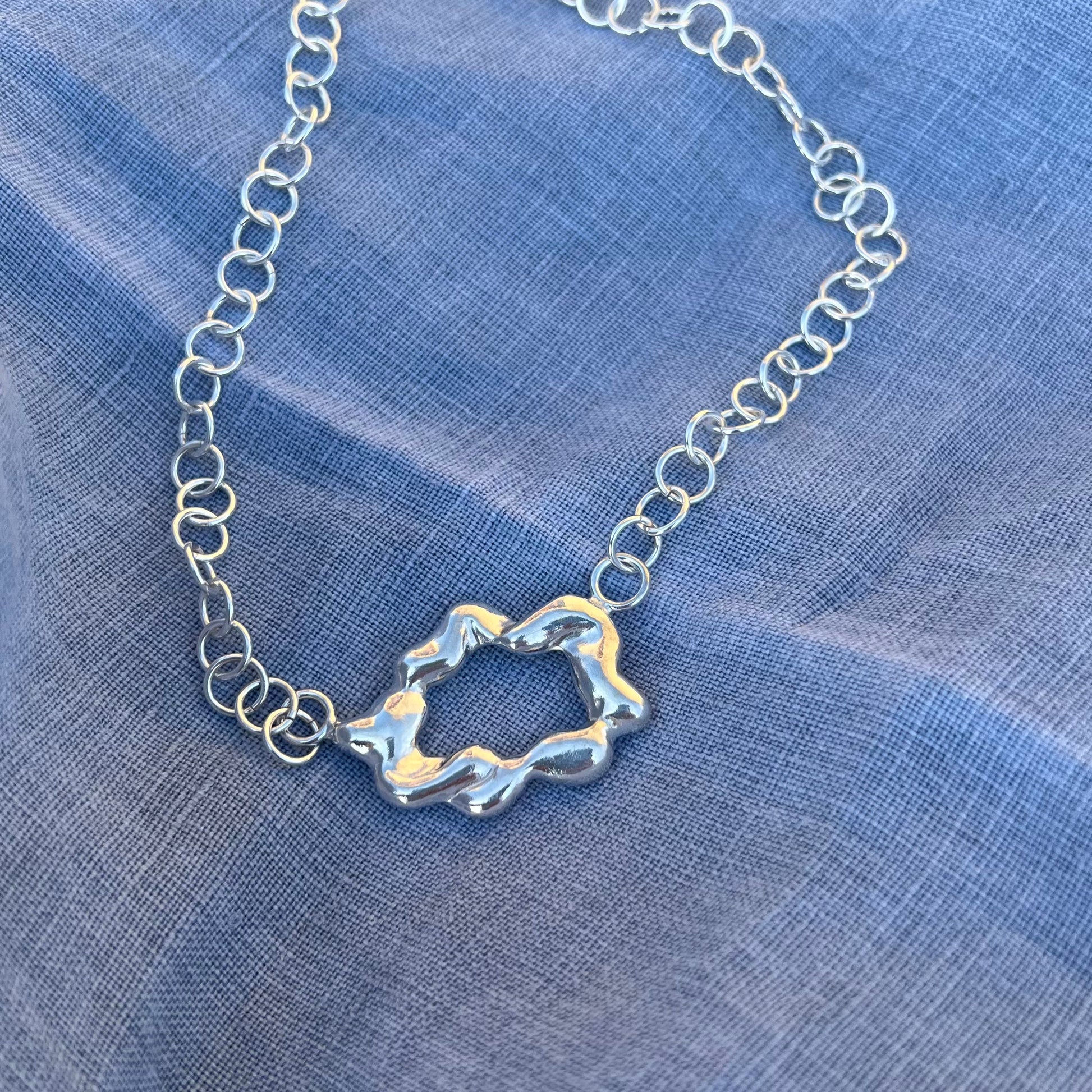 A zoomed in photograph of a chunky, statement handmade sterling silver bracelet on a blue linen background. The centrepiece of the bracelet is shaped like a cloud and it has a handmade hook clasp.