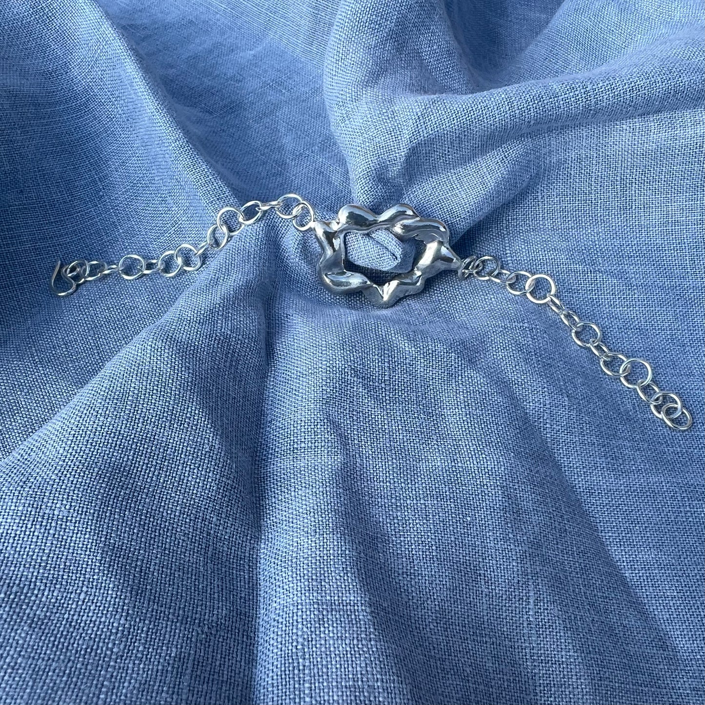 A chunky, statement handmade sterling silver bracelet on a blue linen background. The centrepiece of the bracelet is shaped like a cloud and it has a handmade hook clasp.