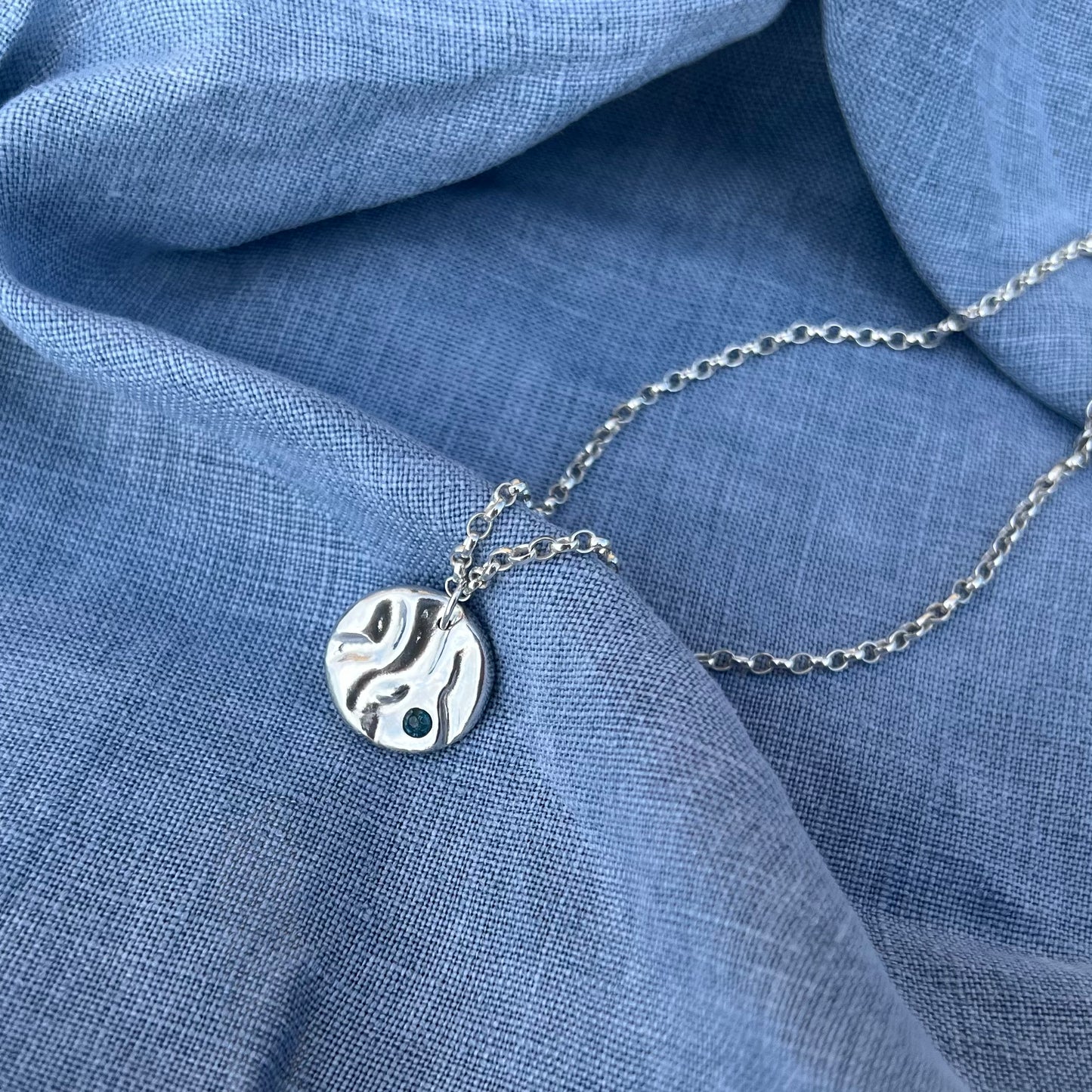 A sterling silver 'Rock-pool pendant" set with a London topaz stone. The pendant is on a belcher chain and the background is of blue linen.