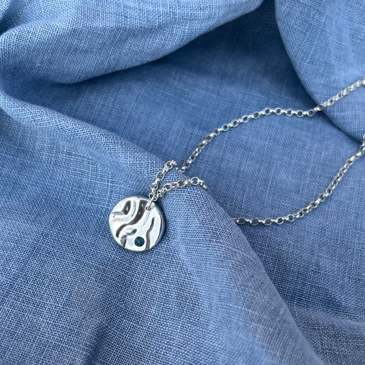 A sterling silver 'Rock-pool pendant" set with a London topaz stone. The pendant is on a belcher chain and the background is of blue linen.