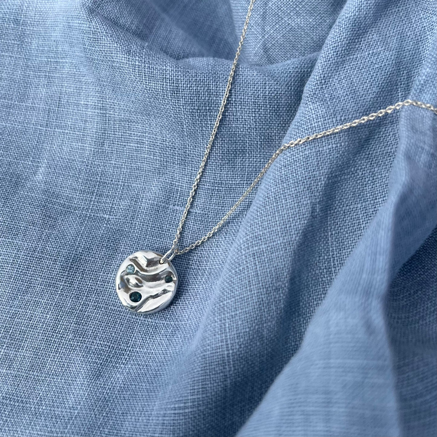 A sterling silver 'Rock-pool pendant" set with two London topaz stones and a sky blue topaz. The pendant is on a belcher chain and the background is of blue linen.
