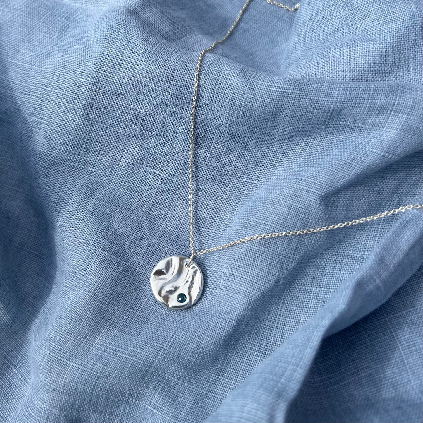 A sterling silver rock pool pendant set with a topaz on a silver chain. The background is blue linen.