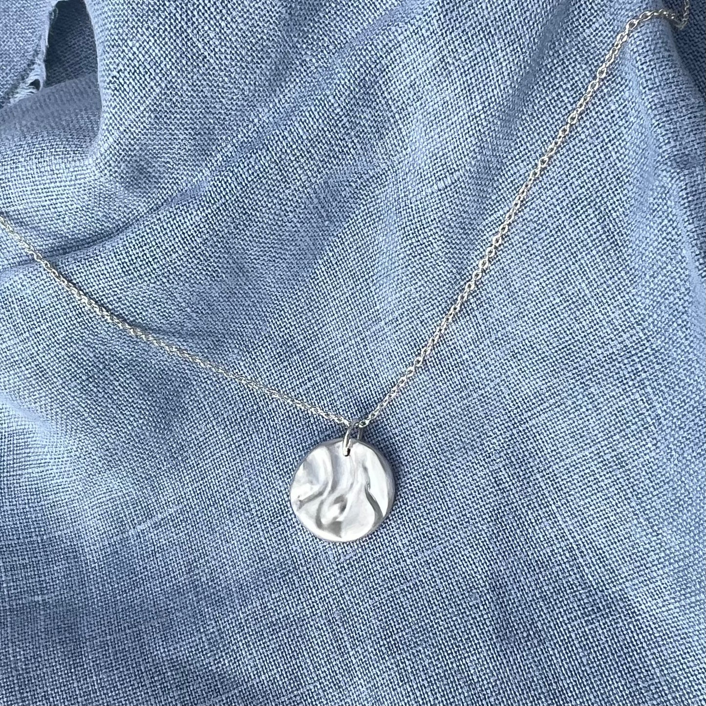 A matt sterling silver 'Rock-pool pendant". The pendant is on a belcher chain and the background is of blue linen.